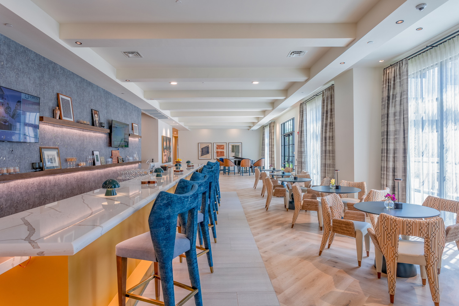 A stylish bar and lounge area featuring a marble countertop, bold blue bar stools, woven rattan seating, and floor-to-ceiling windows at this senior living community in Memphis, TN.