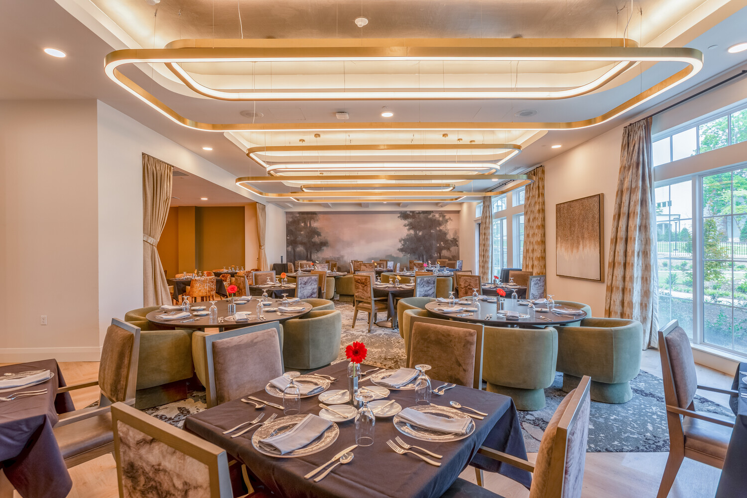 An elegantly set dining room with sage green curved chairs, gold pendant lighting, a scenic mural, and large windows at this senior living community in Memphis, TN, offering a refined and welcoming dining experience.
