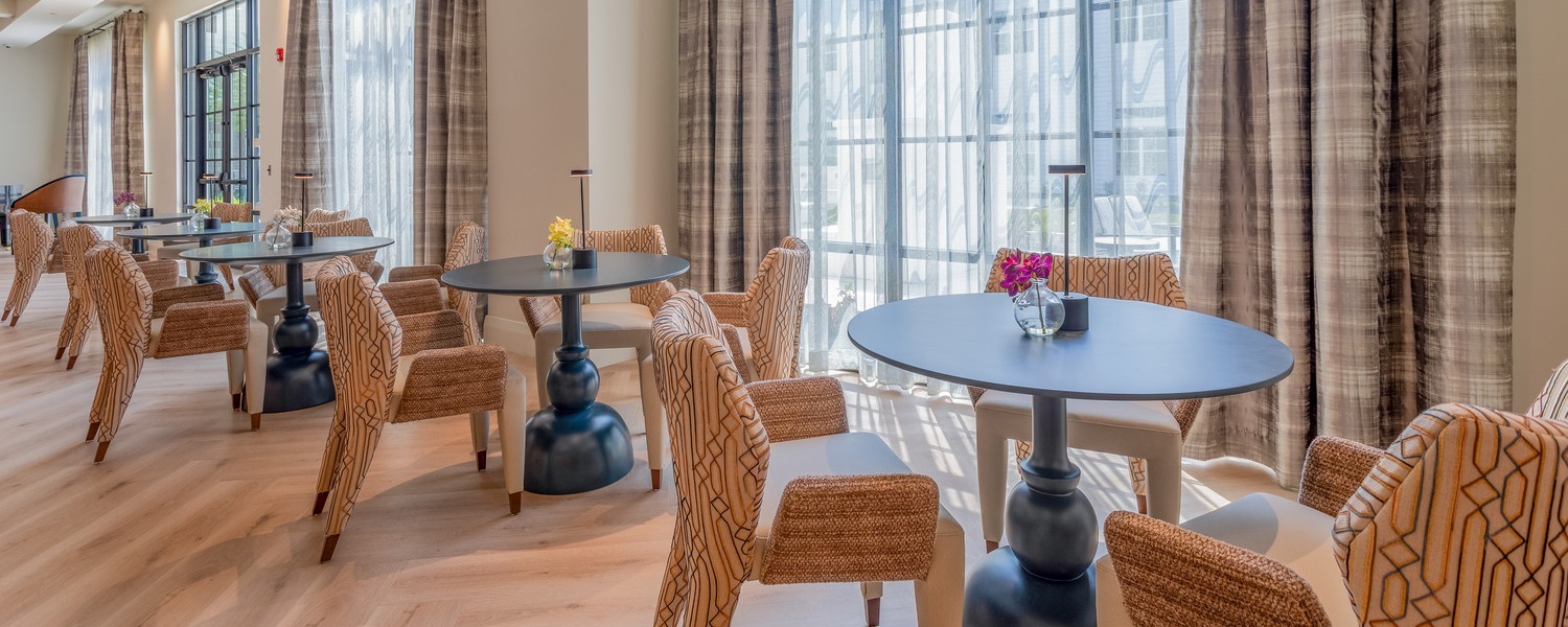 A sunlit lounge area with woven rattan chairs, sleek round tables, and floor-to-ceiling draped windows at this senior living community in Dacula, GA, offering a warm and inviting space to relax.