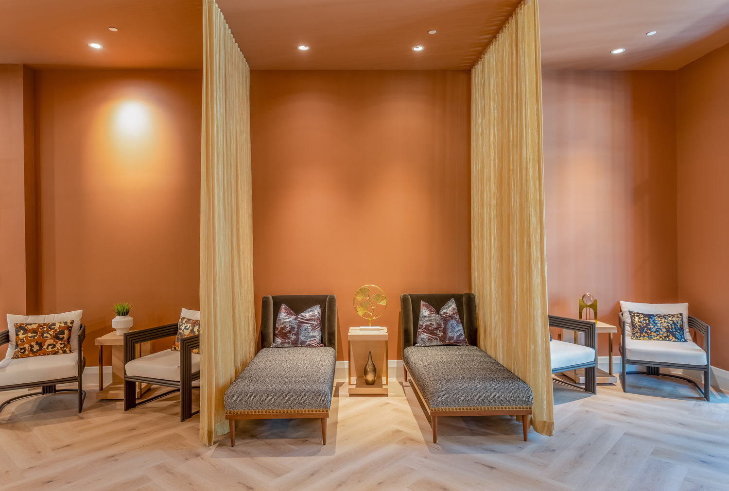A serene spa relaxation room with warm terracotta-colored walls, two upholstered chaise lounges with decorative pillows separated by sheer curtain panels, a small decorative sculpture on a pedestal between them, accent chairs with patterned cushions on either side, and soft recessed lighting above a light herringbone wood floor.