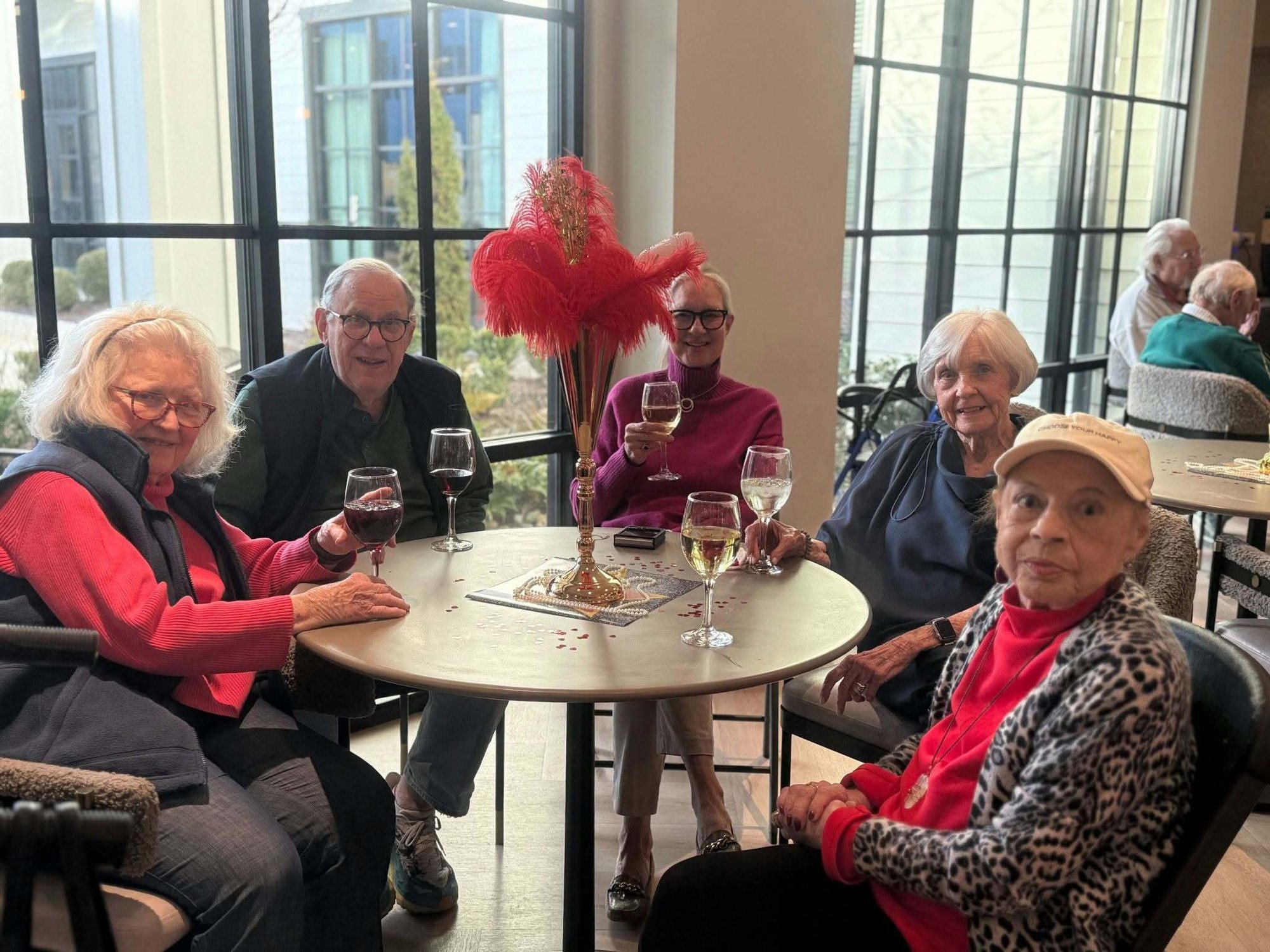 Five people raise wine glasses around a round table adorned with a red feather centerpiece and pearl strands during an Oscars-themed event at this retirement community in Memphis, TN.