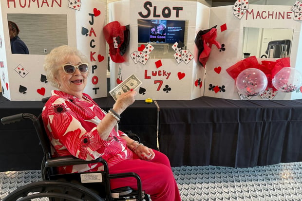 A resident of this assisted living community in Memphis, TN smiles in front of a festive handmade human slot machine display, dressed in red and wearing sunglasses, holding a dollar bill during a themed activity event.