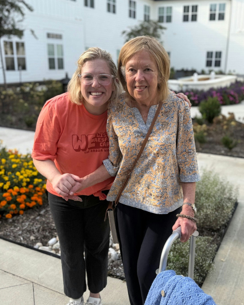 Two women smiling and posing together outdoors in front of a white building with colorful flower beds. The woman on the left wears a coral t-shirt and black pants, while the woman on the right wears a floral blouse and black pants and holds a rolling suitcase.