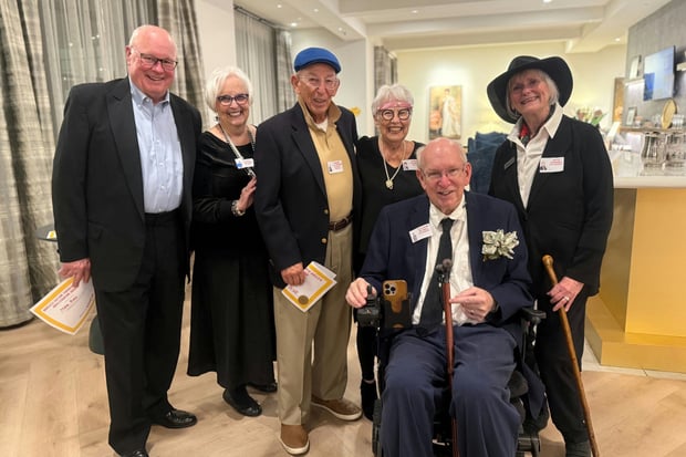 A cheerful group of residents dressed up and smiling together at a themed celebration at Opus East Memphis, a retirement community in East Memphis, TN, with one resident seated in a wheelchair holding an event program.