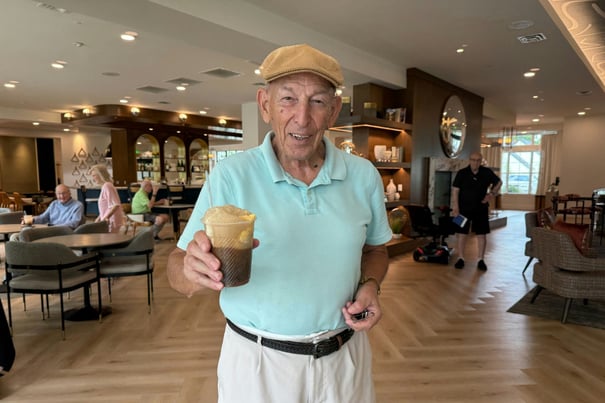 An elderly man wearing a tan cap and light blue polo shirt smiles while holding up an iced coffee drink inside a bright, modern café.