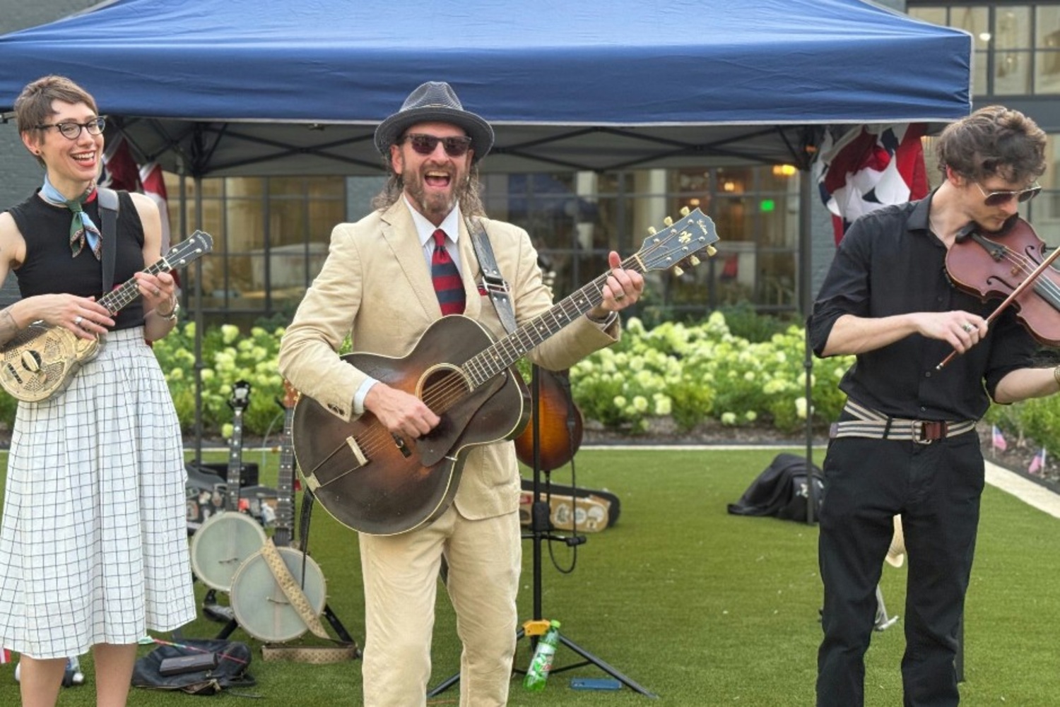 A three-piece band performing outdoors at a community event, with a woman playing a ukulele on the left, a man in a tan suit and fedora playing acoustic guitar in the center, and a younger musician playing violin on the right, with a blue tent and greenery in the background.
