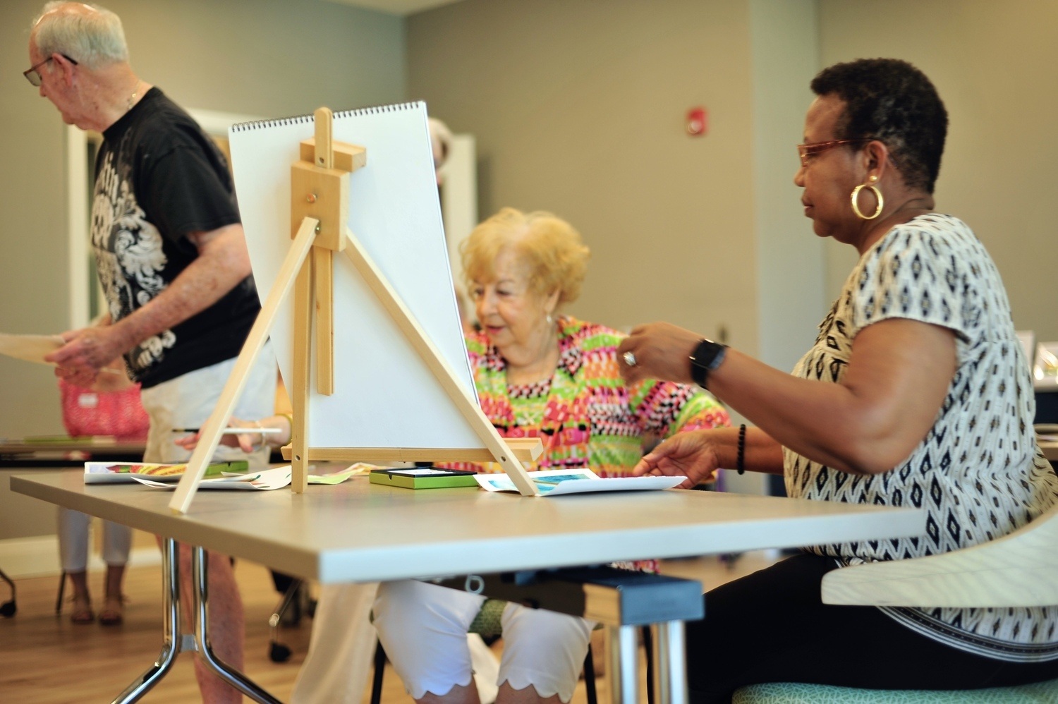 Two older women smile and work together at a table during an art class, with a small canvas on an easel and painting supplies in front of them, while a third resident stands in the background in a bright, modern activity room
