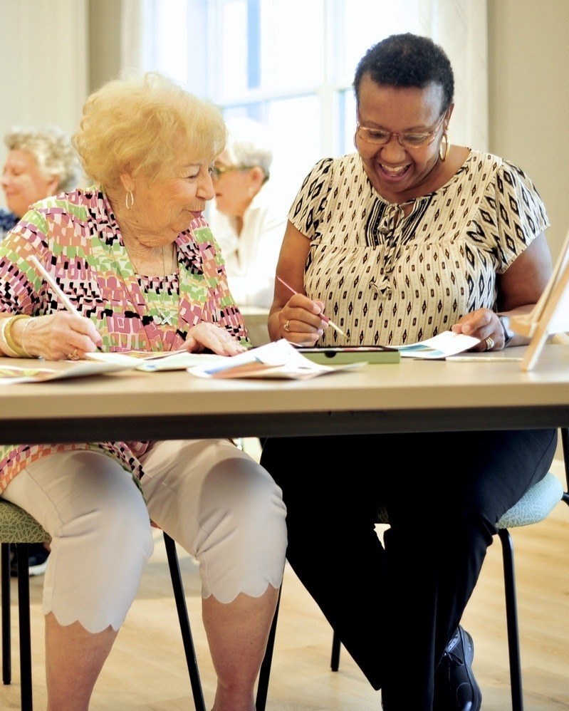 A woman in a colorful top laughs warmly while sharing artwork during a group activity at Opus East Memphis, reflecting the engaging senior lifestyle Memphis TN residents enjoy.