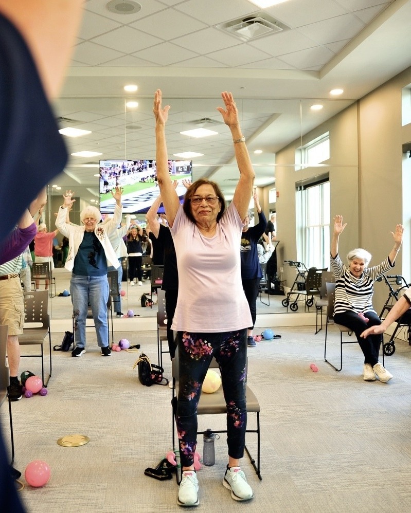 A group of older adults participating in a fitness class in a bright community room, with one woman in the foreground raising both arms overhead in an exercise movement, surrounded by other residents and colorful exercise equipment.