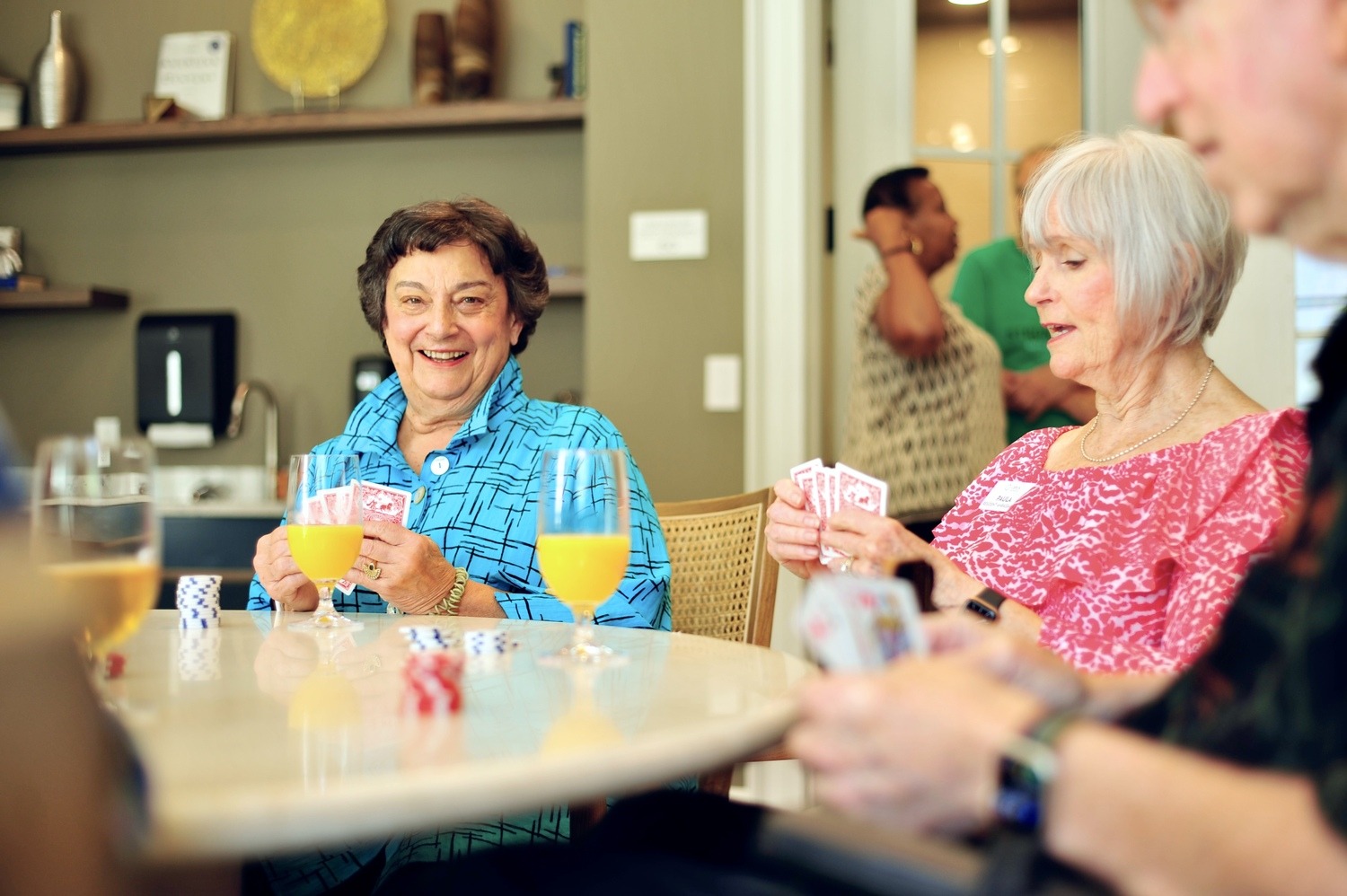 Two older women laugh and smile while playing cards at a table with glasses of orange juice, joined by a third player in the foreground, in a warm and casual common room setting with other residents visible in the background.