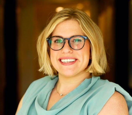 A staff member at Opus East Memphis senior living in East Memphis, TN, smiling warmly in a teal blouse and glasses, photographed in a softly lit hallway.