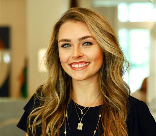 A staff member at Opus East Memphis, a senior living community in East Memphis, TN, smiling brightly in a black top with layered gold necklaces, photographed in a warmly lit interior.