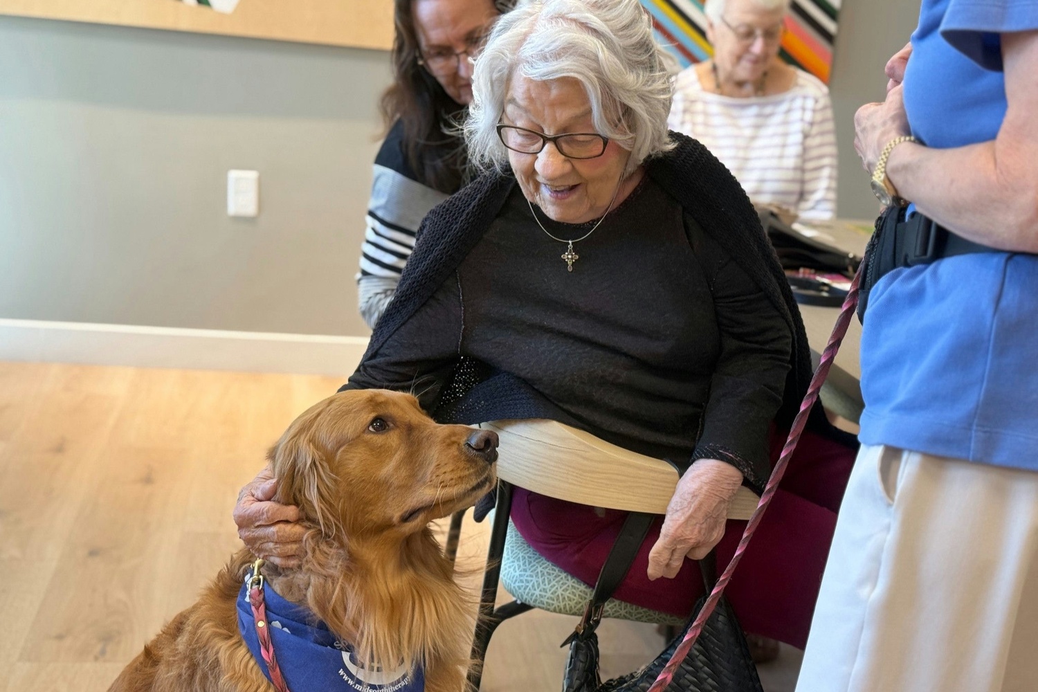An older woman seated in a wheelchair smiles as she pets a golden retriever therapy dog wearing a blue vest, while a caregiver and other residents look on in a bright, modern common room with light hardwood floors.