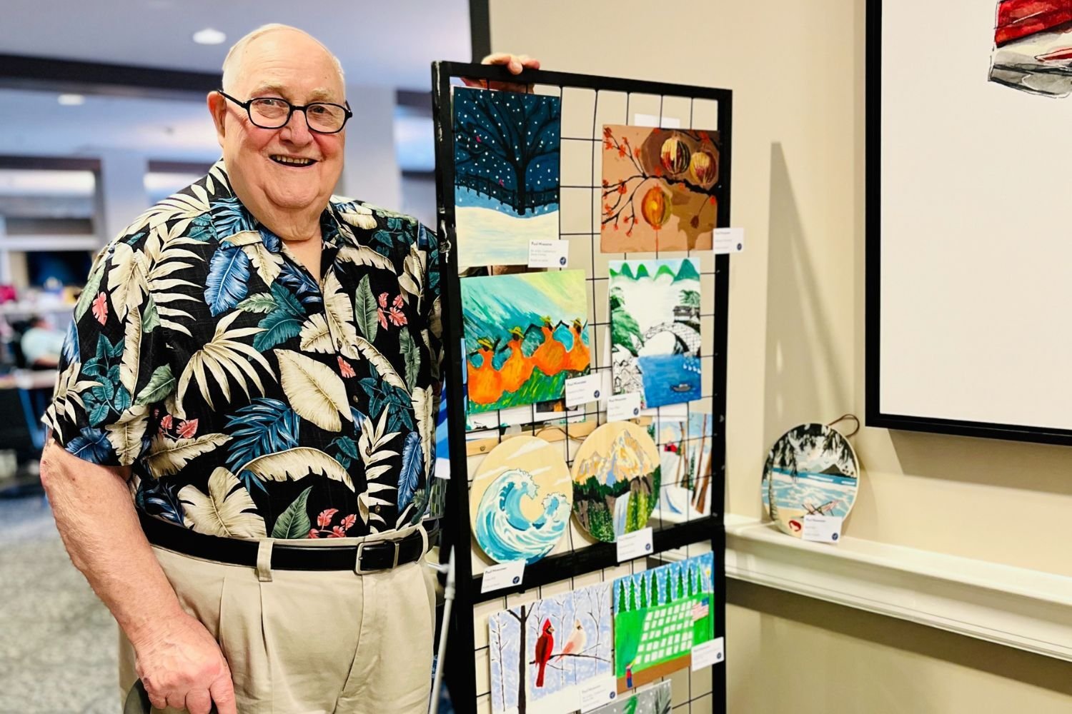 man in a Hawaiian shirt stands next to his art with a big smile on his face