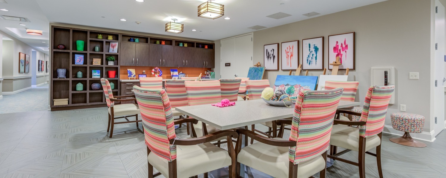 A bright activity room with striped upholstered chairs, a wooden table, colorful storage bins, and framed abstract art, showcasing the vibrant common spaces found in this 55+ senior living Woodbridge and resort-style senior living Woodbridge community.