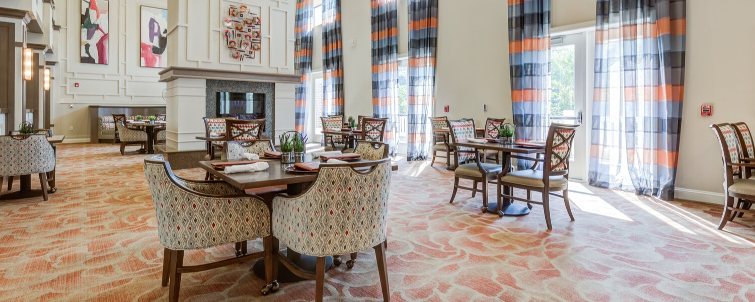 A bright dining lounge with patterned area rugs, a fireplace, colorful artwork, and tall windows framed by orange and blue curtains, reflecting the resort-style senior living Woodbridge charm of this senior living Woodbridge VA community.