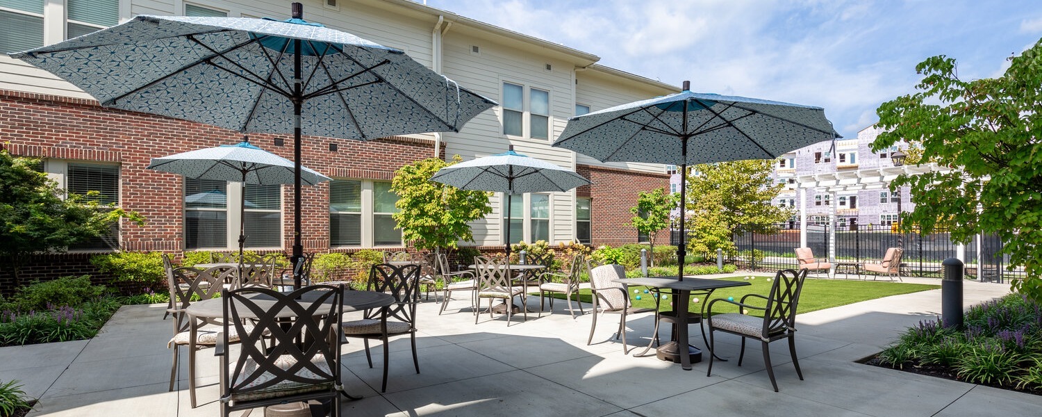 A sunny outdoor patio with blue umbrellas, black metal tables and chairs, and landscaped greenery beside the brick community building, reflecting the resort-style senior living Woodbridge charm and outdoor comfort of senior living Woodbridge VA.