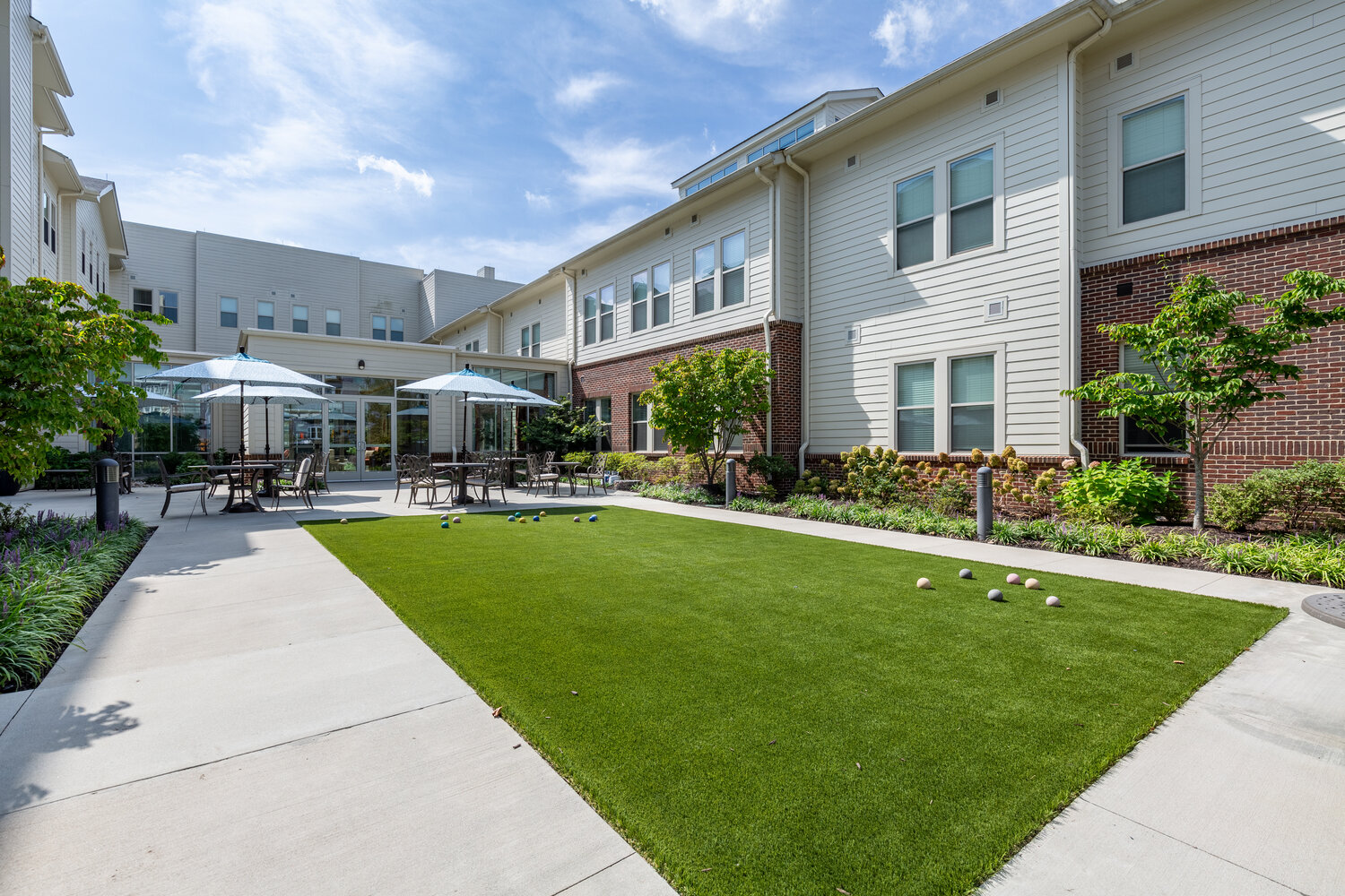 A landscaped courtyard with a manicured lawn, patio seating, and umbrellas invites residents outdoors, reflecting the nature-access senior living Woodbridge setting and convenience of senior living near Potomac Mills.
