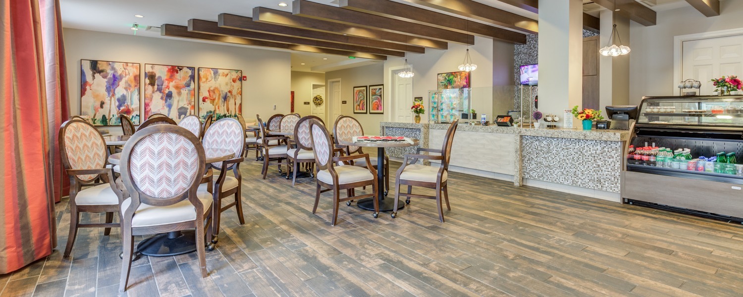 Cozy dining area at Arbor Terrace senior living Woodbridge VA, featuring wood beam ceilings, round tables with patterned chairs, warm wood-look flooring, and a beverage counter, reflecting the resort-style senior living Woodbridge atmosphere residents enjoy.