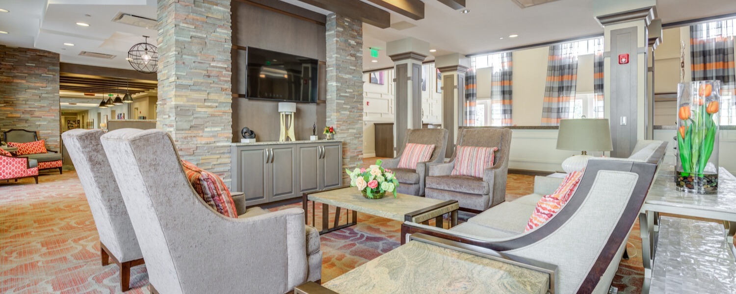 Elegant lounge area featuring a stone accent wall, mounted TV, patterned armchairs, and a coffee table with fresh flowers, showcasing the resort-style senior living Woodbridge ambiance and inviting comfort of senior living Woodbridge VA.