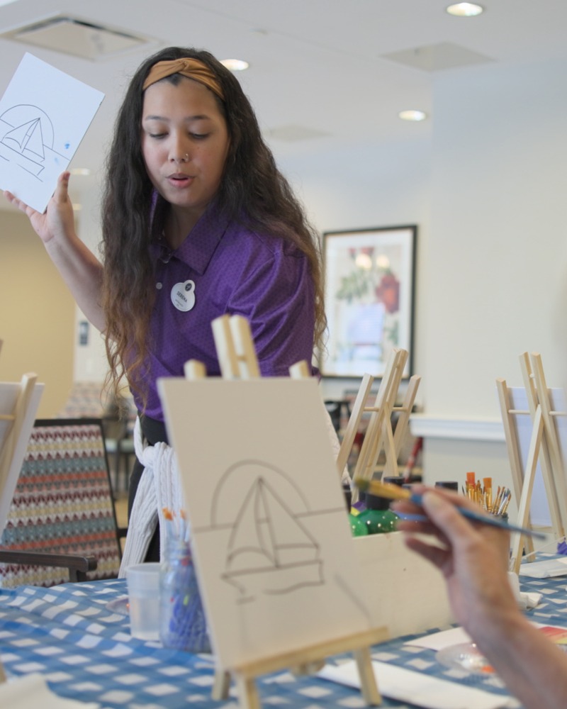 A staff member holds up a sailboat sketch while guiding residents through a painting class at easels, reflecting the creative arts programming within this active senior community Prince William County and senior living Woodbridge VA.