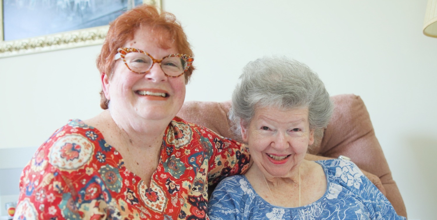 Two women share a warm embrace and joyful smile while relaxing together on a cozy couch, reflecting a heartfelt visit and the welcoming spirit of senior living near me, part of the vibrant senior living Manassas VA community connections at Arbor Terrace.