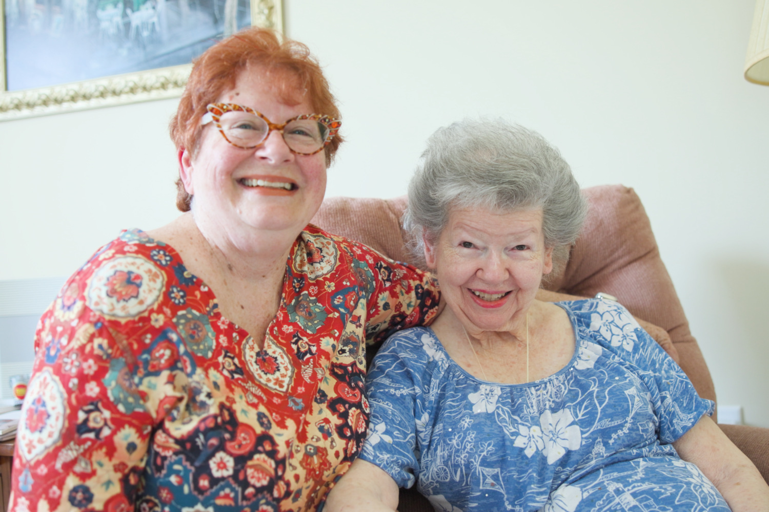 Two women share a warm embrace and big smiles while relaxing together on a cozy sofa, reflecting the welcoming friendships found within senior living Woodbridge VA and this wellness-focused senior living Northern Virginia community.