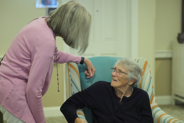 A staff member leans in warmly to chat with a seated resident in a bright striped armchair, capturing a caring moment within this wellness-focused senior living Northern Virginia community near Prince William Forest Park.