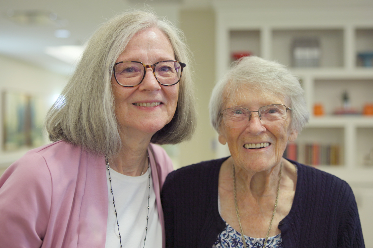 Two smiling women pose closely together indoors near a bookshelf, sharing a warm friendship that reflects the welcoming community connections found in this active senior community Prince William County and senior living Woodbridge VA.