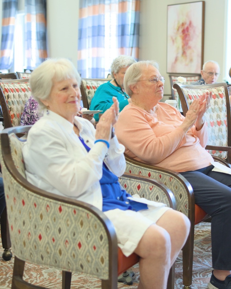 Residents clap and smile together during a lively event in a bright common area with patterned armchairs, capturing the joyful community spirit of this active senior community Prince William County and senior living Woodbridge VA.