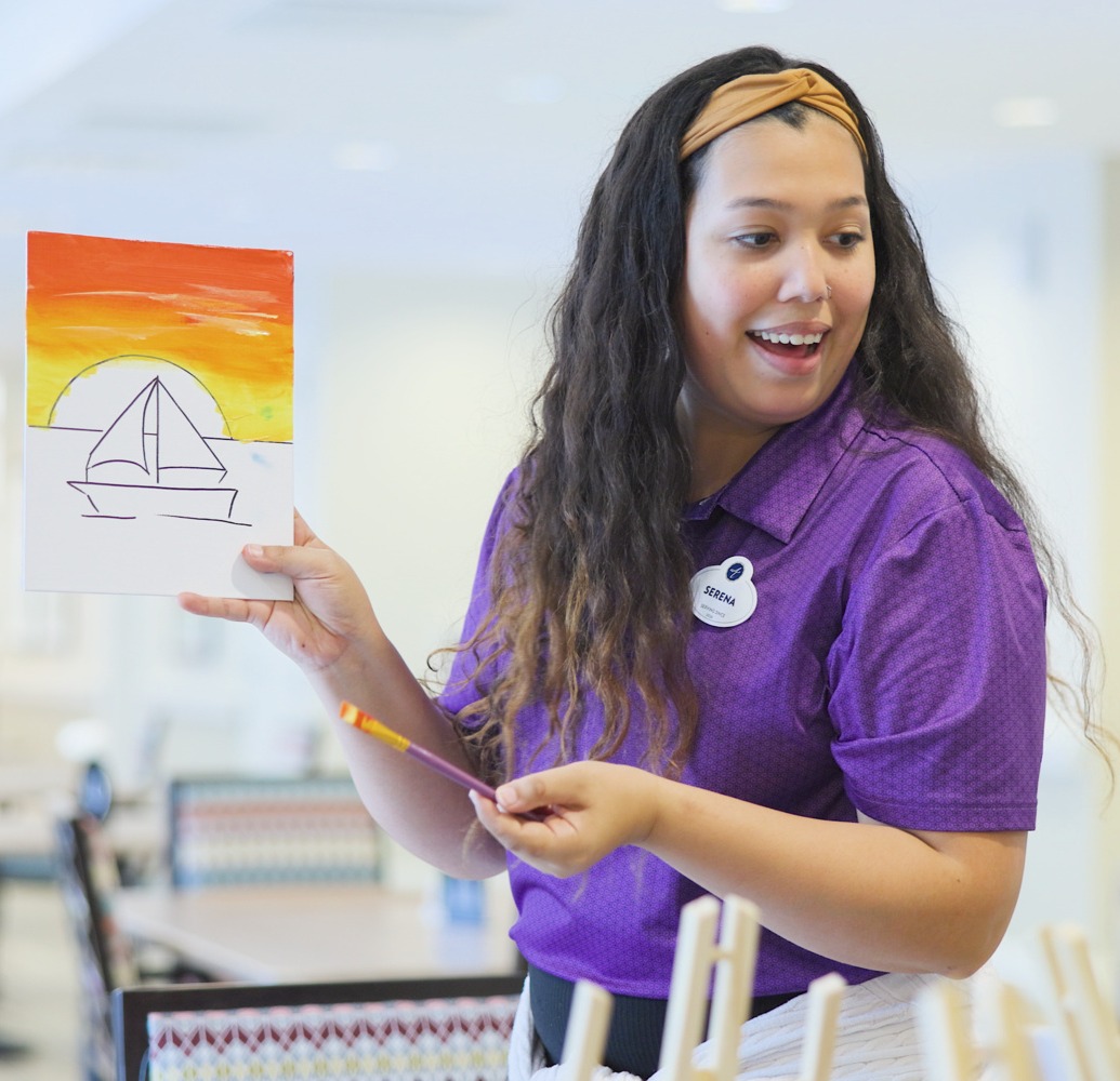 A staff member smiles while holding up a colorful sailboat sunset painting created during an art class, reflecting the creative programming within this active senior community Prince William County and senior living Woodbridge VA.