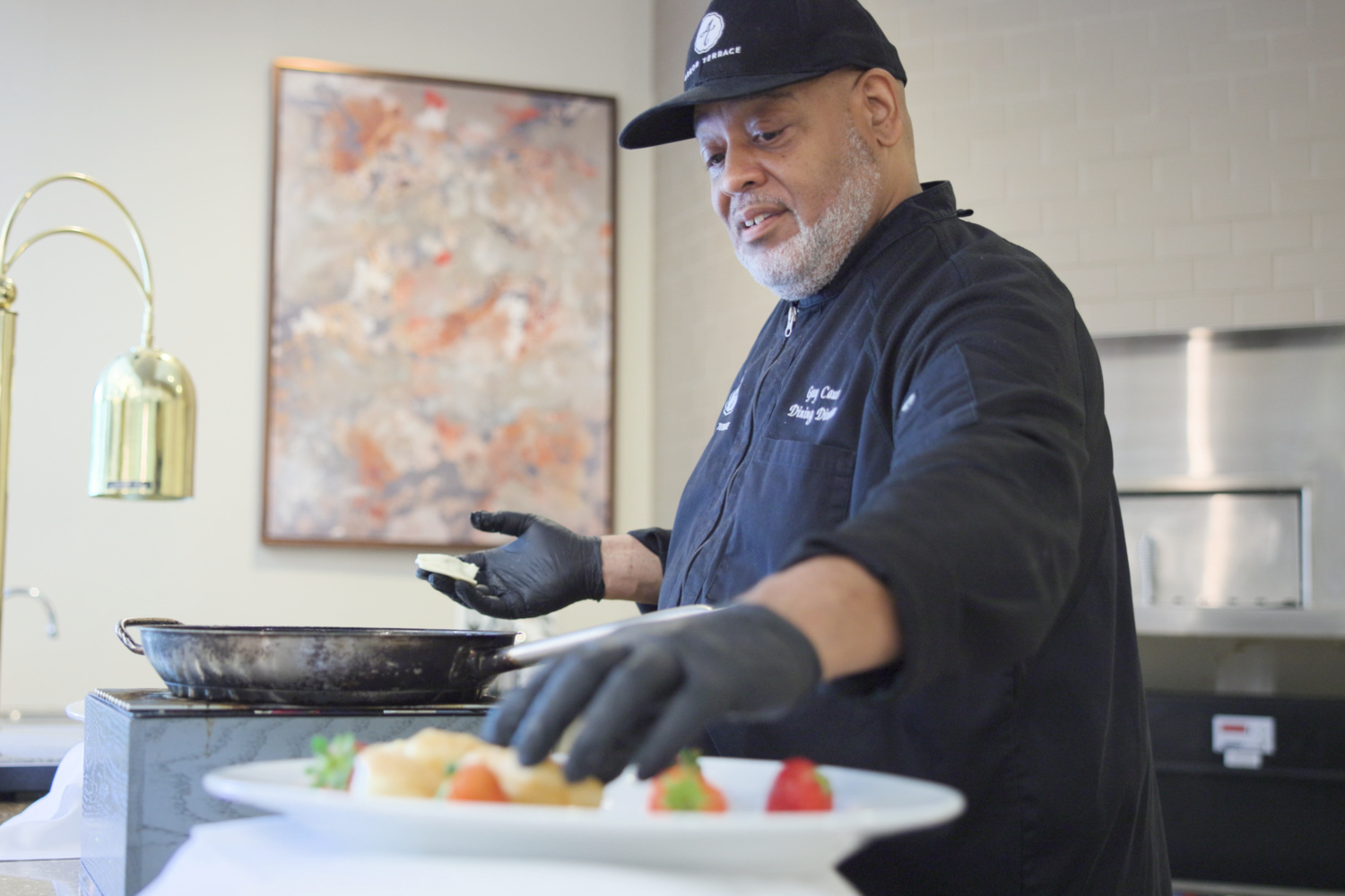 A chef in a black uniform and cap carefully plates a fresh dish garnished with strawberries and herbs in a modern kitchen, reflecting the award-winning dining found at this senior living Woodbridge VA and resort-style senior living Woodbridge community.