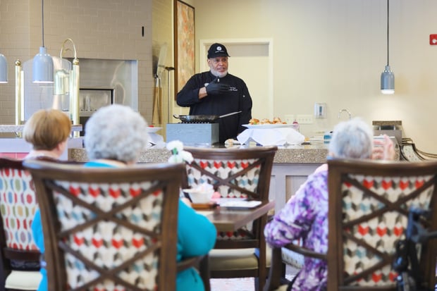 A chef prepares a dish tableside for residents gathered for Sunday brunch, capturing the warm dining tradition central to senior living near Potomac Mills within this active senior community Prince William County.