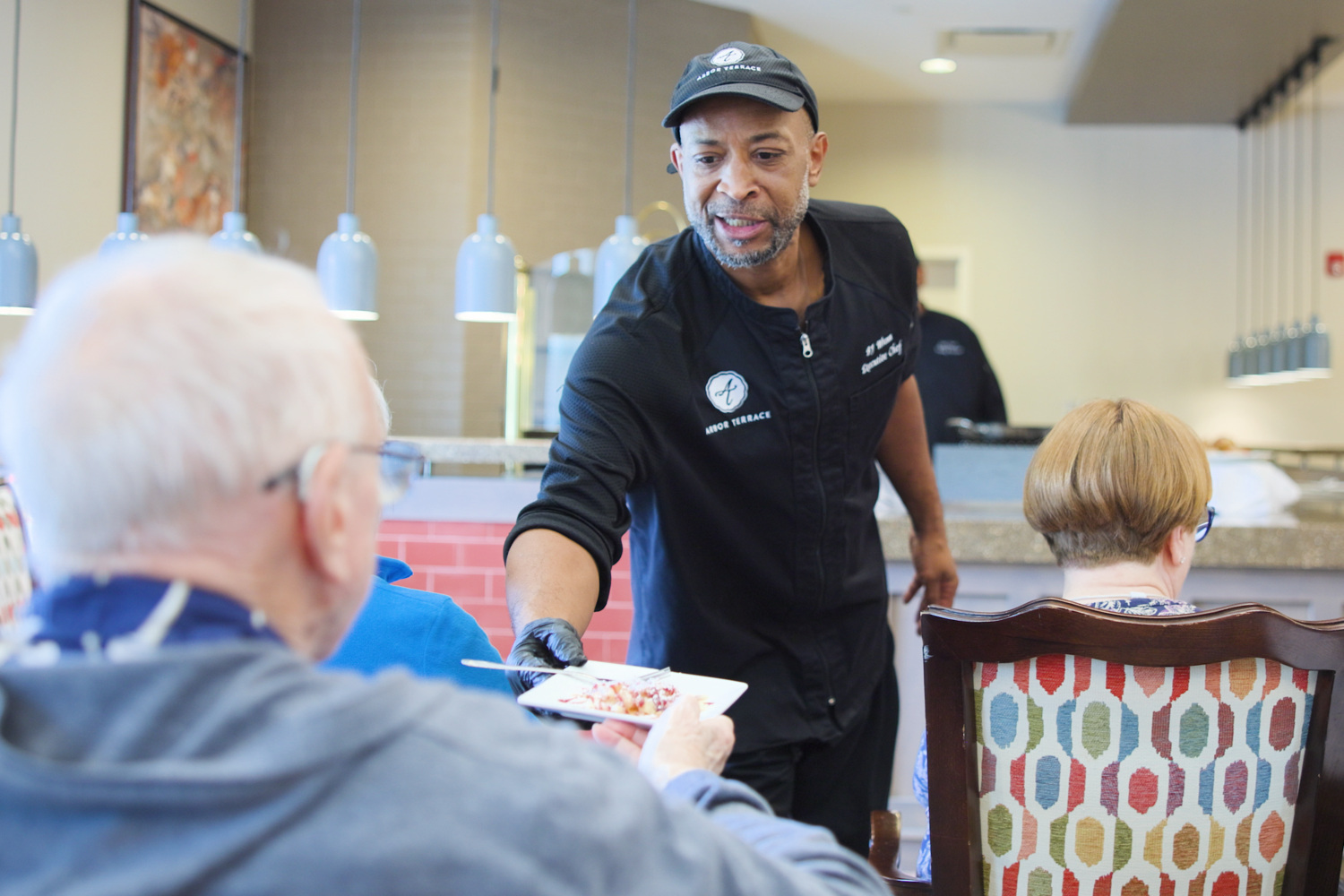 A chef serves a beautifully plated dish to residents seated at a colorful dining table, capturing the warm hospitality of this active senior community Prince William County and senior living near Potomac Mills.