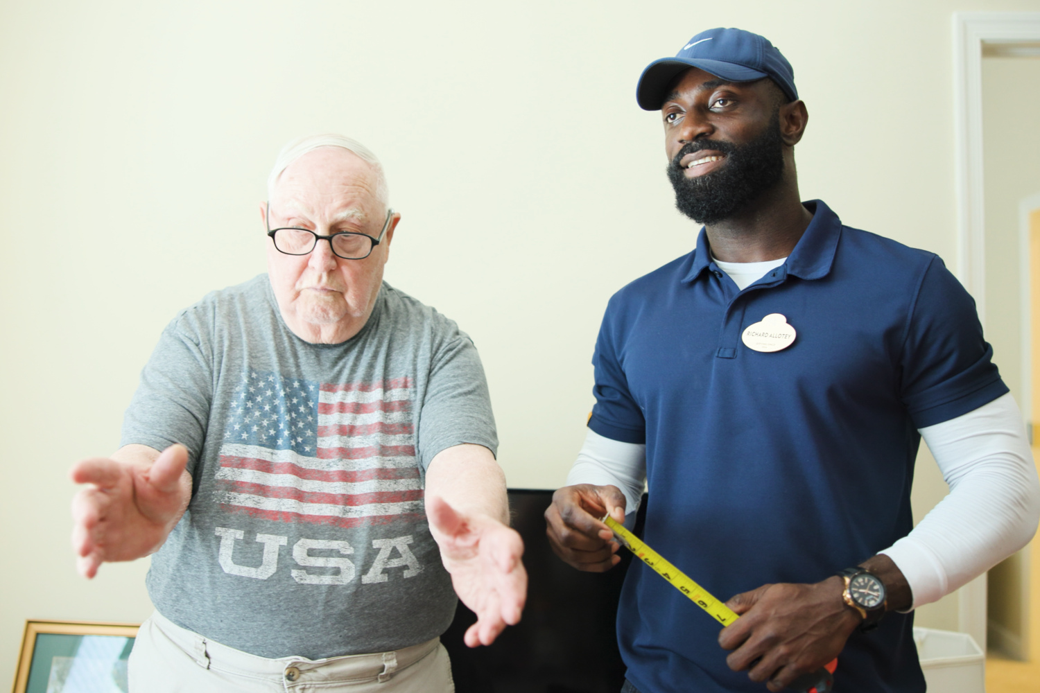 A resident reaches out playfully while a staff member measures with a tape measure, sharing a lighthearted moment that reflects the fun, active senior community Prince William County lifestyle within this 55+ senior living Woodbridge community.