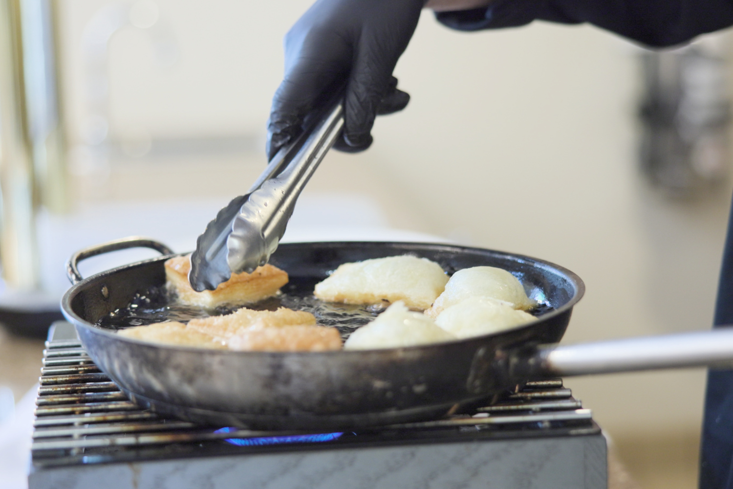 A chef's gloved hand uses tongs to flip golden bread in a sizzling skillet over an open flame, capturing the fresh, chef-prepared meals central to dining at this senior living Woodbridge VA community.