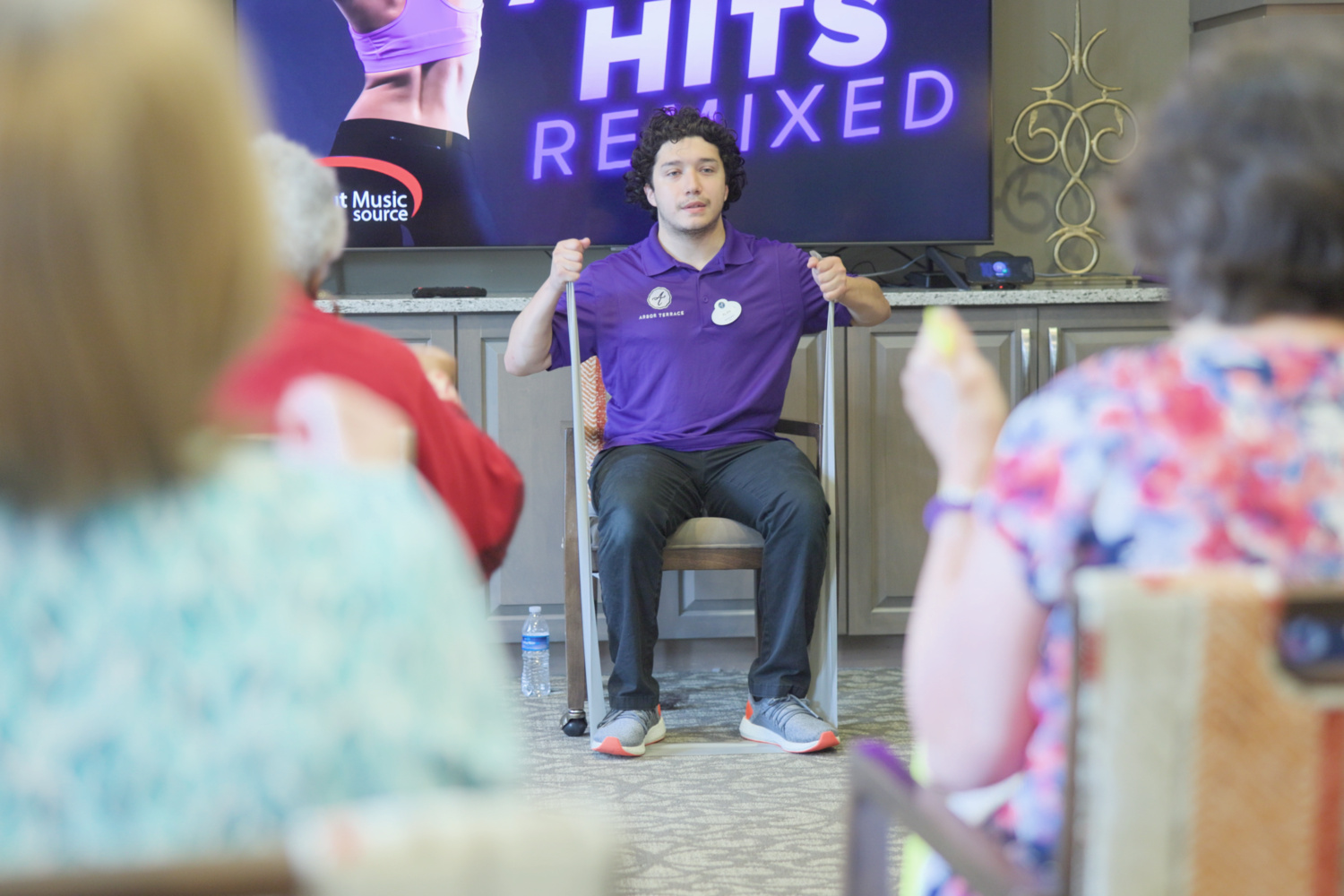 A team member in a purple Arbor Terrace polo leads a seated fitness class with a resistance band as participants follow along in front of a large screen at Arbor Terrace Prince William Commons.