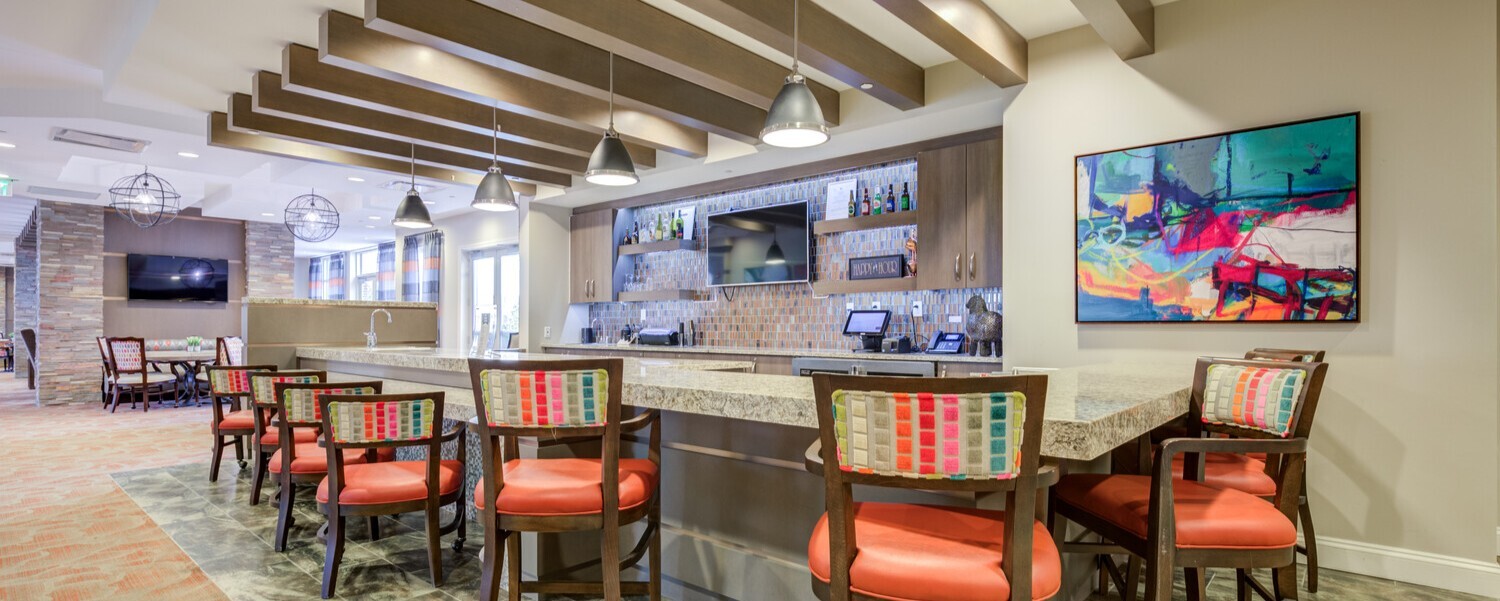 Stylish onsite bar and lounge at Arbor Terrace Prince William Commons featuring a granite countertop, colorful patterned barstools, wall art, and a wood beamed ceiling, part of the vibrant amenities within this senior living community in Woodbridge VA.
