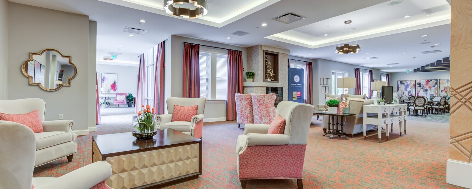 Spacious living room at Arbor Terrace Prince William Commons with cream and coral wingback chairs, a wood coffee table, a decorative fireplace, and layered lighting, reflecting active senior living in Woodbridge VA.