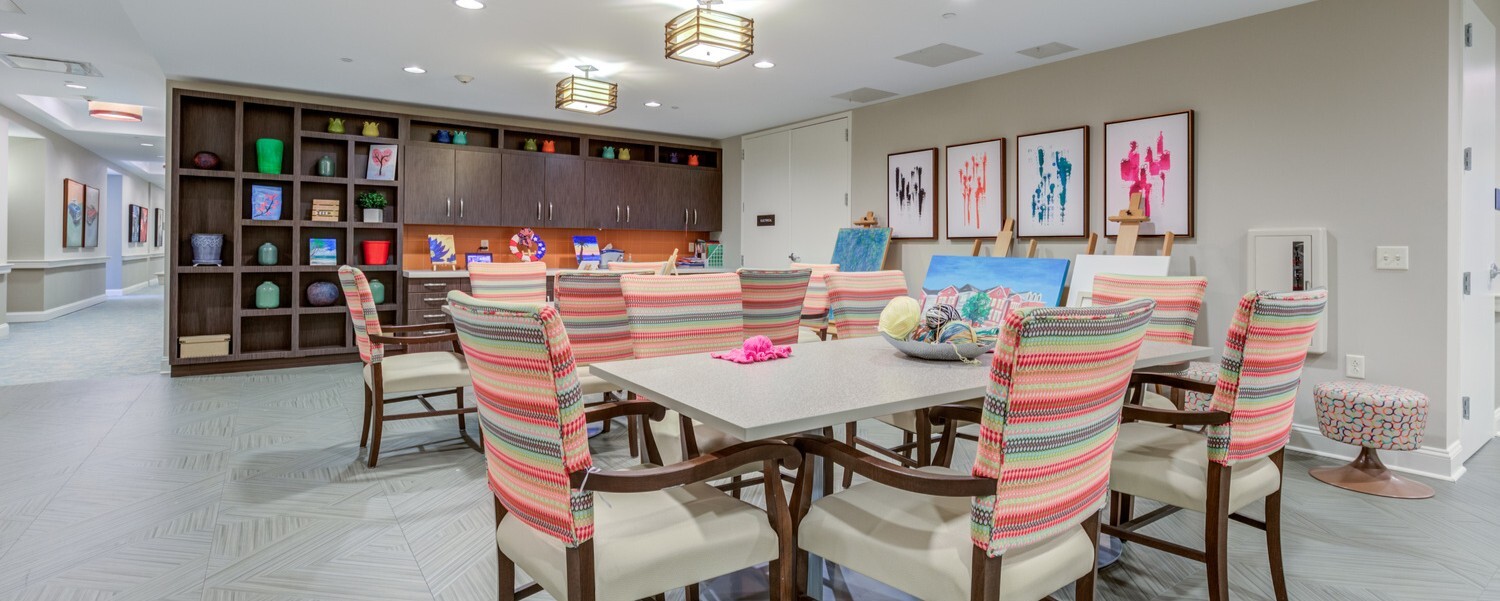 Bright creative arts space at Arbor Terrace Prince William Commons with a long communal table, striped upholstered chairs, display shelving, and easels with framed artwork, part of an active senior community in Woodbridge VA.