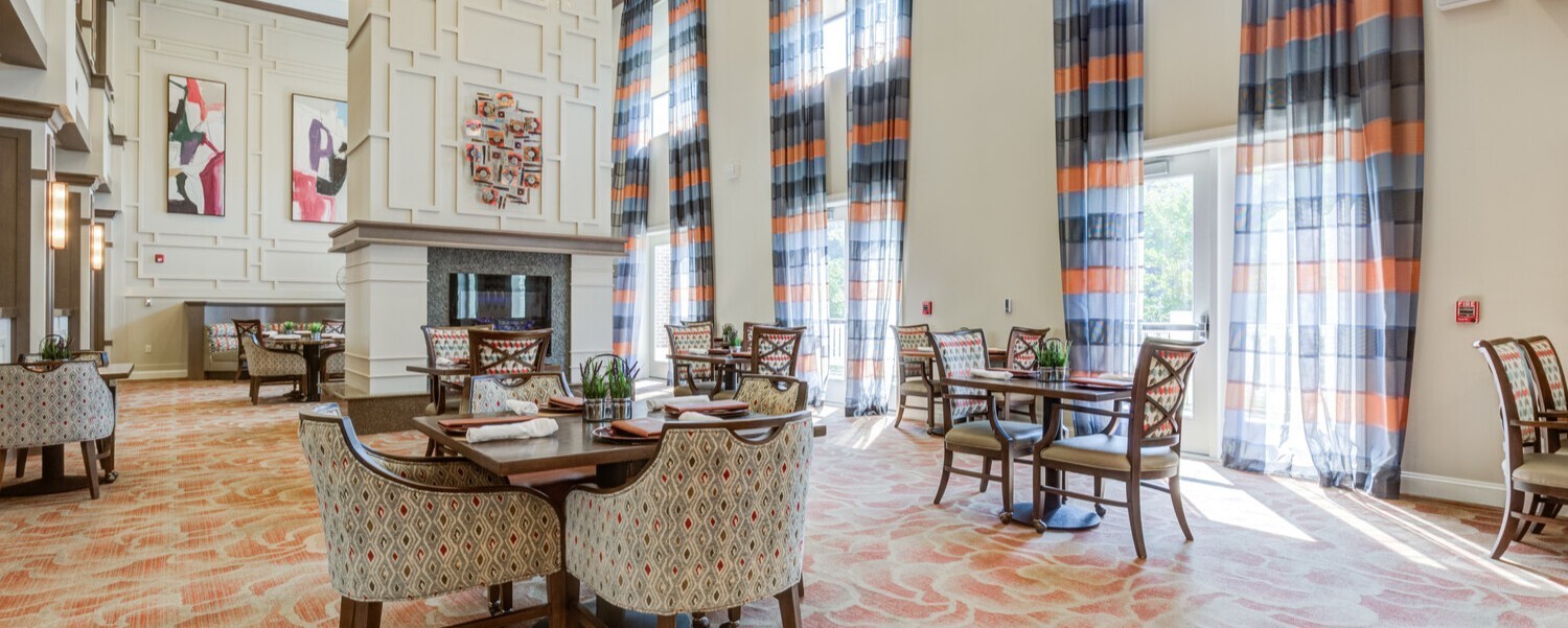Sunlit dining room at Arbor Terrace Prince William Commons with floor-to-ceiling striped drapery, a stone fireplace, patterned wingback chairs, and bistro-style tables, part of senior living in Woodbridge VA.