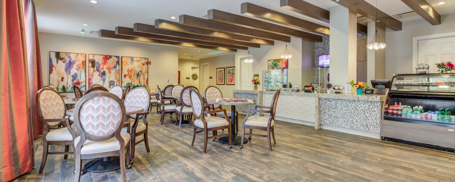 Charming café and bistro at Arbor Terrace Prince William Commons with wood beamed ceilings, oval-back chairs, colorful wall art, and a mosaic-tiled service counter with a bakery display, part of senior living in Woodbridge VA.