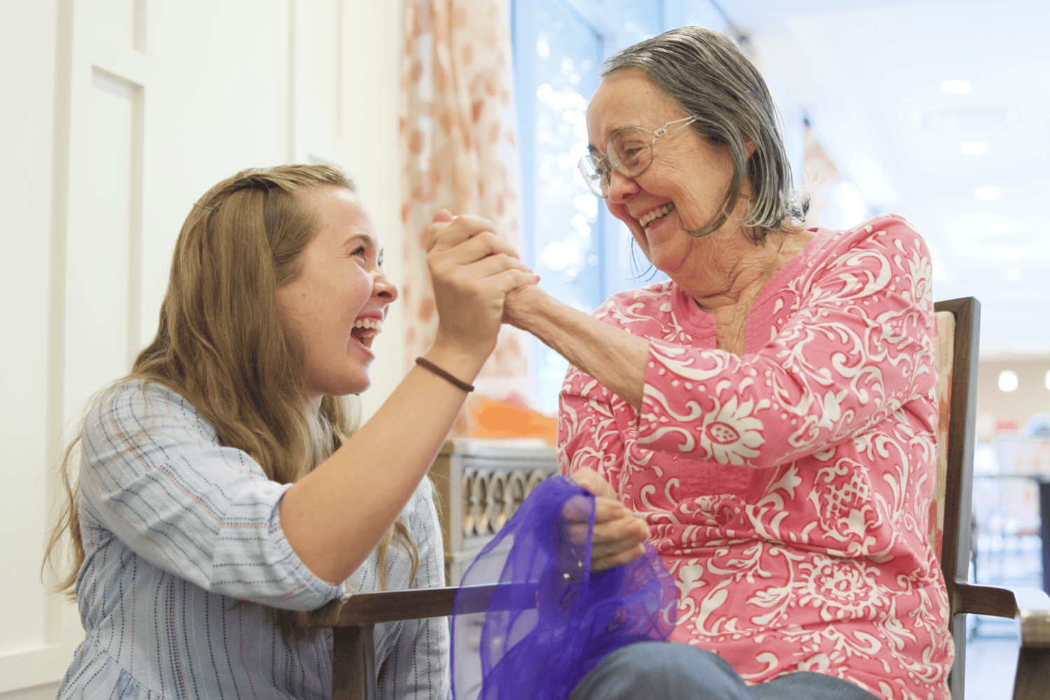 A kneeling young woman and a woman in a pink floral top holding a bright purple scarf laugh together as they clasp hands in a sunlit room at Arbor Terrace Prince William Commons.
