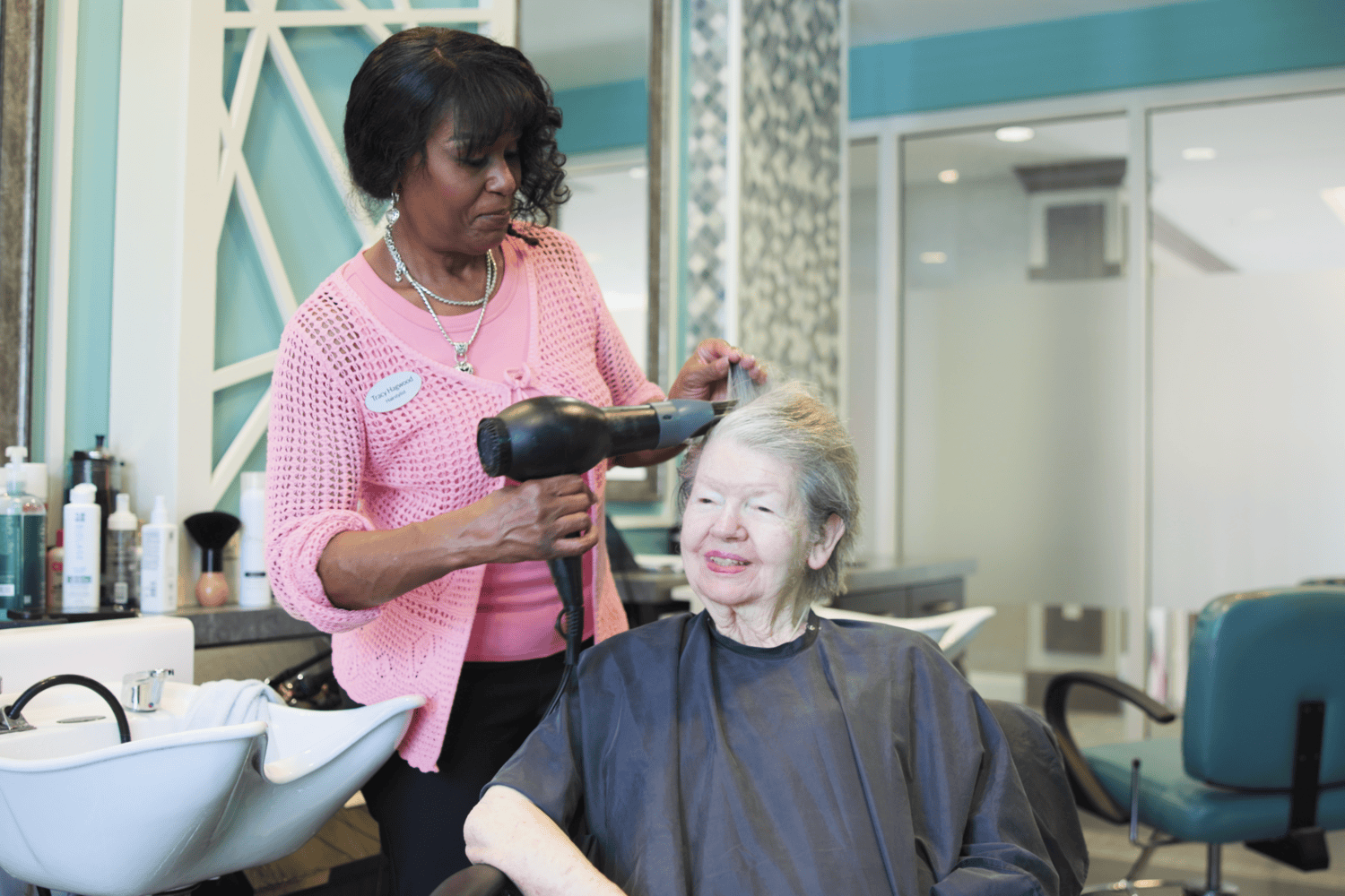 A stylist in a pink crochet cardigan and silver necklace blow-dries the hair of a smiling woman seated beside a white shampoo bowl in the bright aqua salon at Arbor Terrace Prince William Commons.