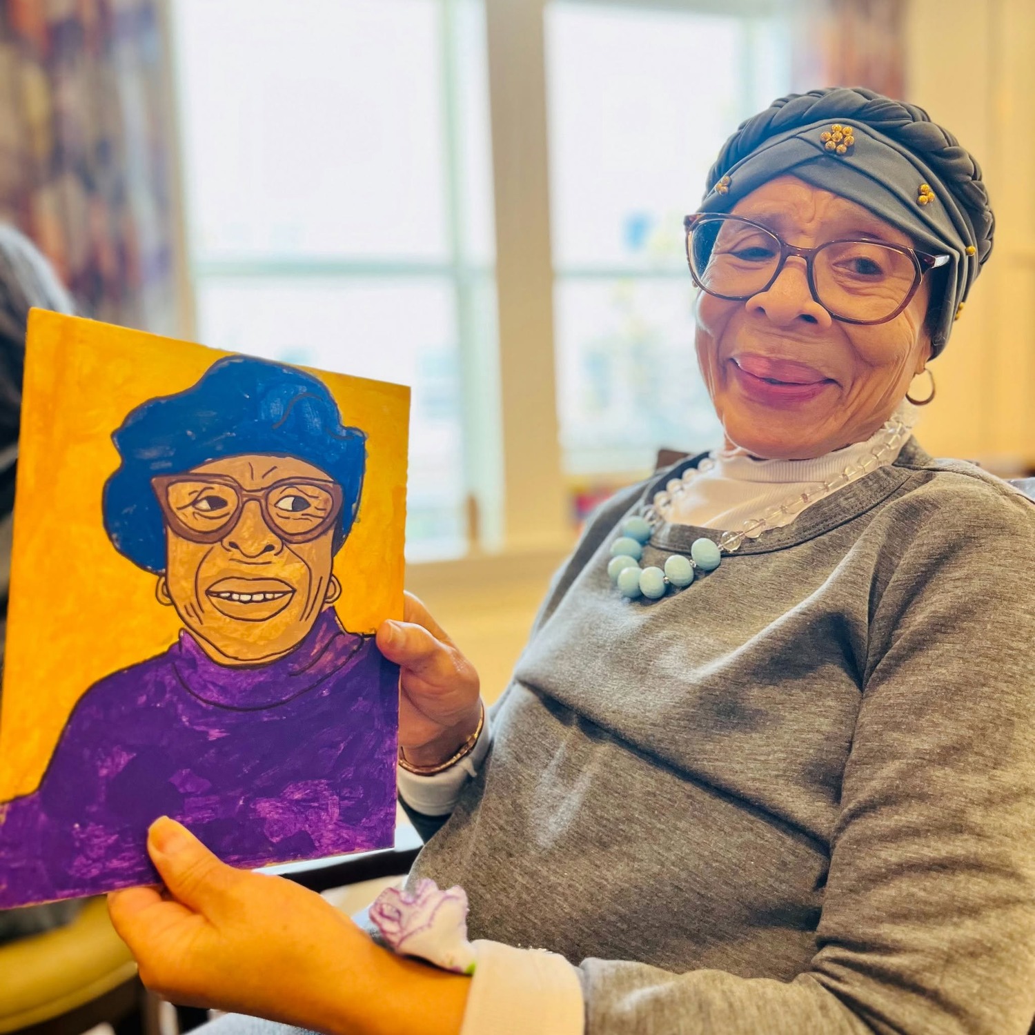 A resident proudly holds up a vibrant self-portrait painting she created, smiling warmly by a sunlit window, capturing the creative spirit celebrated within this active senior community Prince William County and senior living Woodbridge VA.