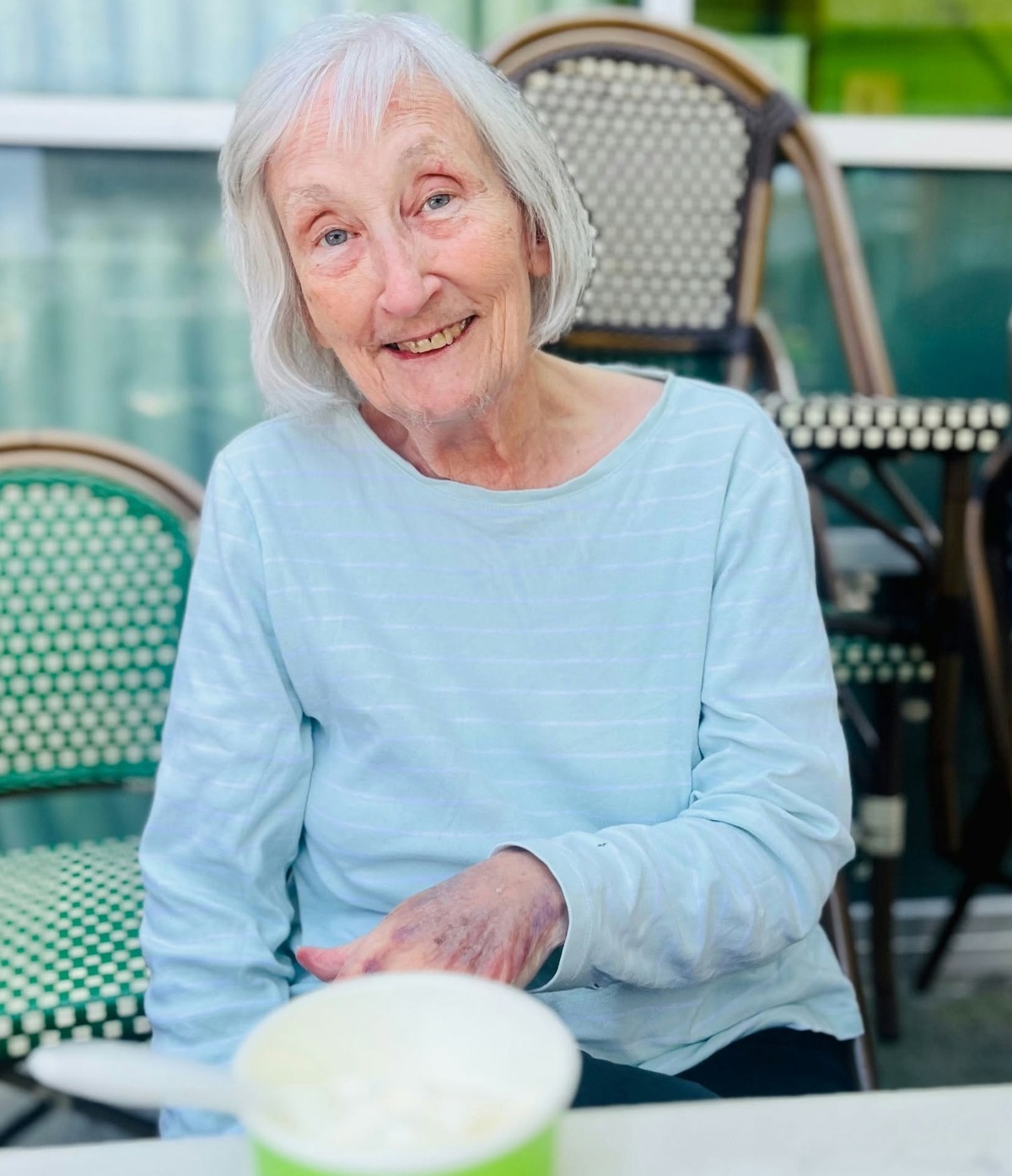A resident smiles warmly while seated at an outdoor cafe table with wire-back chairs, enjoying a cool drink in the sunshine, reflecting the resort-style senior living Woodbridge outdoor lifestyle at senior living Woodbridge VA.