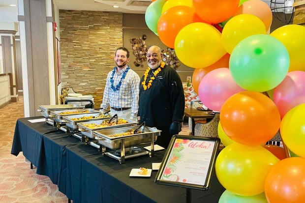 Two staff members smile behind a catering table topped with chafing dishes and colorful balloon decor, celebrating a festive gathering that reflects the vibrant spirit of senior living near Potomac Mills and senior living Occoquan VA.