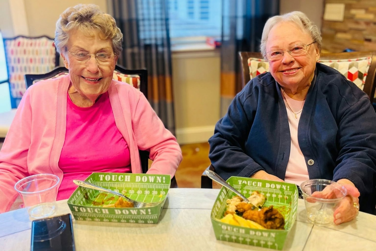 Two residents enjoy a game-day snack together at a cozy table, smiling over football-themed treat boxes and drinks, capturing the fun community spirit of senior living Woodbridge VA and this active senior community Prince William County.