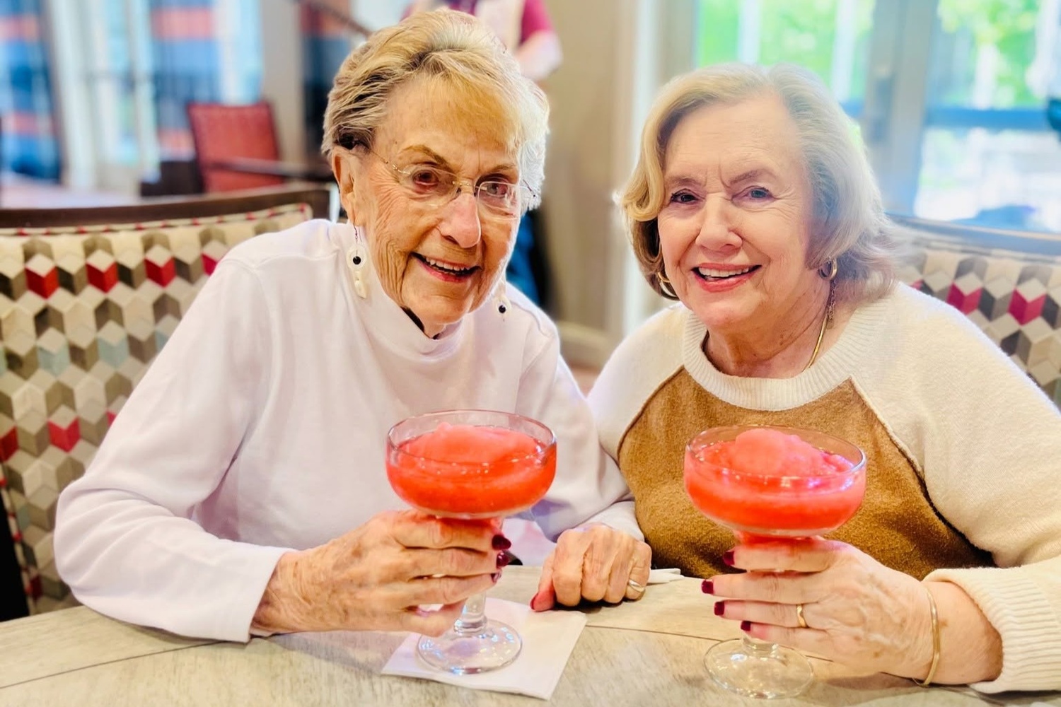 Two residents raise colorful frozen drinks in a cheerful toast, celebrating together in a bright dining space that reflects the resort-style senior living Woodbridge atmosphere and wellness-focused senior living Northern Virginia lifestyle.