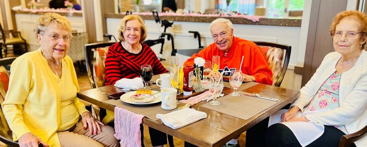 Four residents gather around a table for a festive meal, laughing and enjoying drinks together, reflecting the resort-style senior living Woodbridge hospitality found in this senior community near Prince William Forest Park.
